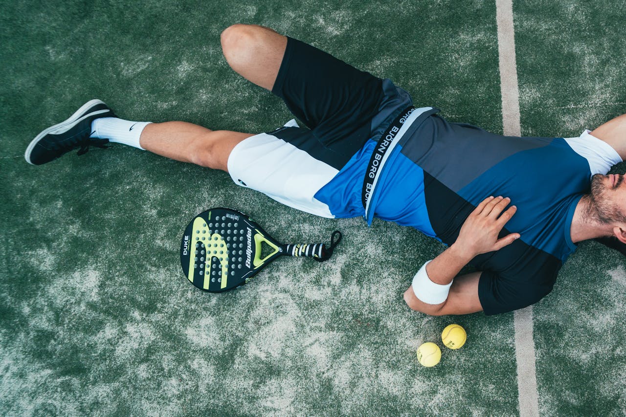 Tired young athlete resting on padel court with racket and balls nearby.